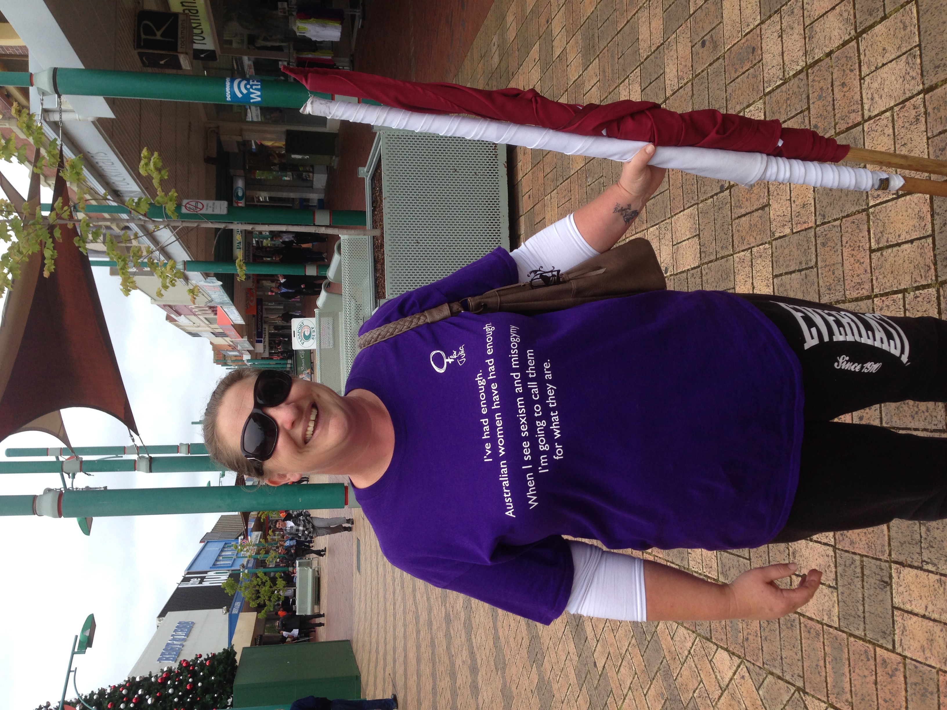 Alison Butcher in Rooke Street Mall Devonport, smiling, wearing sunglasses and a purple t-shirt that reads 'I've had enough. Australian women have had enough. When I see sexism and misogyny I'm going to call them for what they are.'.